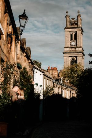 Historic stone church tower and row houses in Bath, England with moody sky and traditional street lamp casting shadows.