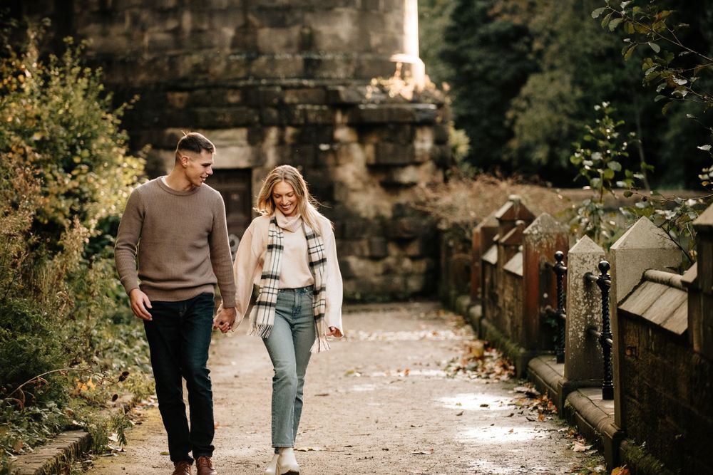 A cozy autumn couple walks together on a stone path bordered by rustic architecture and greenery in natural light.
