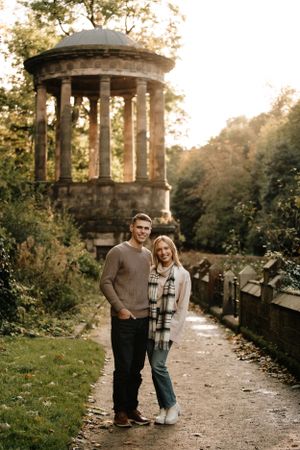 A couple stands on a path near a classical stone gazebo in a romantic autumn garden setting at sunset.