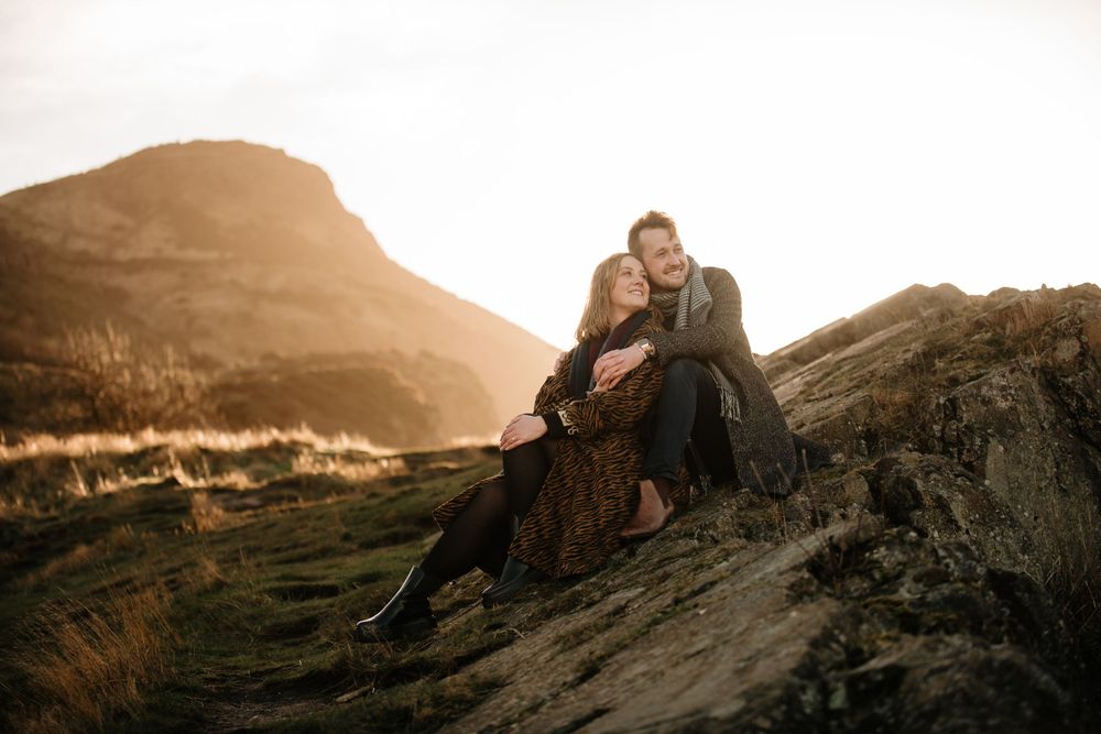 A couple sits close together on rocky terrain at sunset with golden mountains in the background.