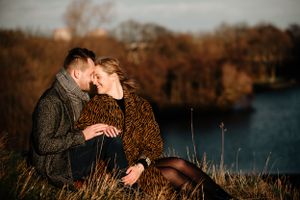A couple shares an intimate moment at sunset by a lake while sitting in golden autumn grass.