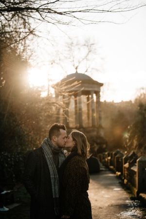 A couple shares a romantic moment on a path with a gazebo and golden sunset light filtering through bare winter trees.