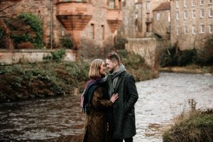 A couple embraces near a river with brick buildings in the background on an overcast day.