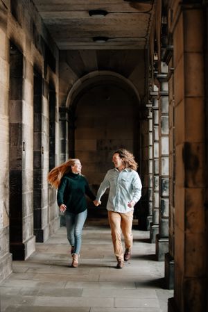 Two people run joyfully through a stone archway corridor with sunlight streaming in from the left side.