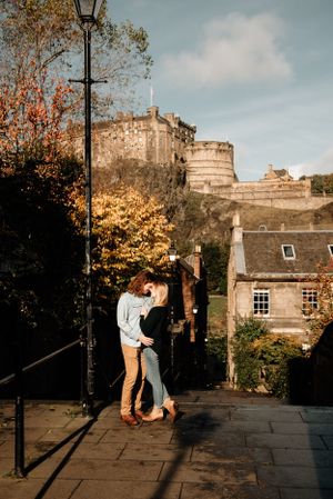A couple embraces on a stone path with Edinburgh Castle and autumn trees visible in the backdrop during golden hour.