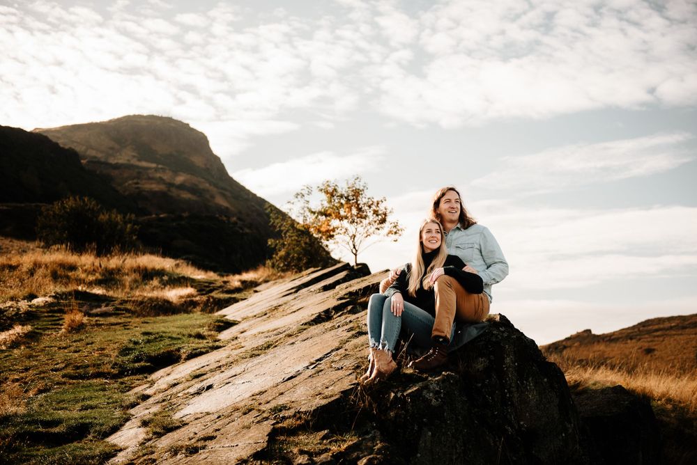 Two people sit together on a rocky mountain overlook during golden hour with scenic mountain views in the background.