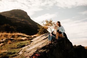 A couple sits together on a large rock formation at sunset with mountains and autumn foliage in the background.
