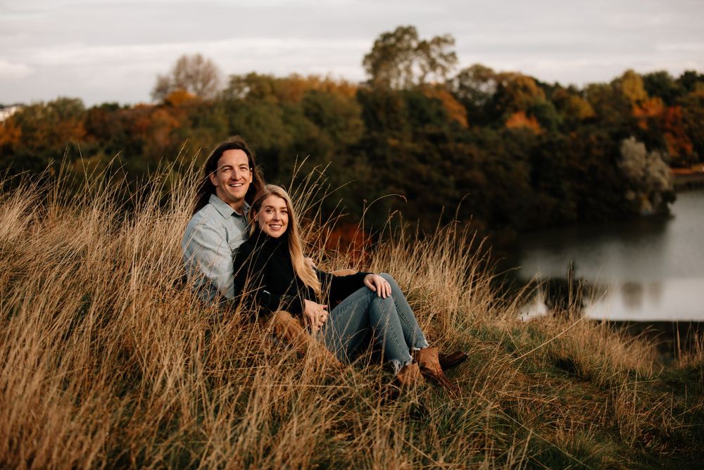 Two people sit together on grassy hill overlooking peaceful lake during golden autumn afternoon.