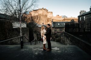 A romantic couple stands on a historic stone walkway in Edinburgh with the castle and old town buildings in the background.