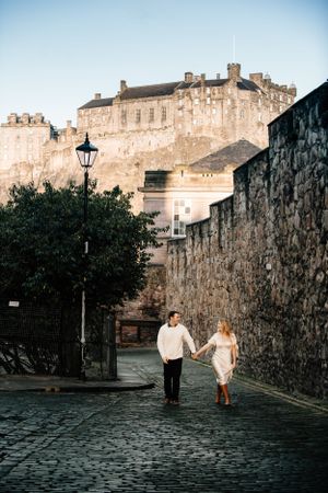 A couple walks along a cobblestone street beneath Edinburgh Castle on a romantic evening in Scotland.