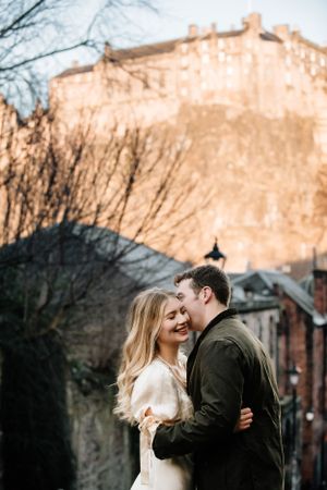 A romantic couple embraces during a winter photoshoot with Edinburgh Castle looming in the background at sunset.