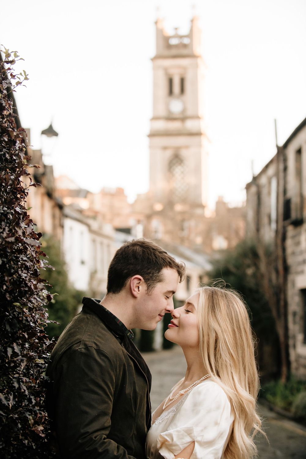 A romantic couple share an intimate moment on a cobblestone street with a historic church tower in the background.