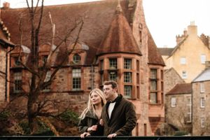 A couple stands together near historic brick buildings with turrets and ornate Victorian architecture in Scotland.