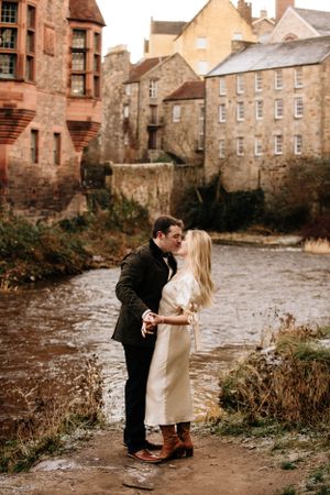 A romantic couple embraces along a riverside path with historic stone buildings and architecture in the background.