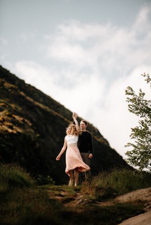 Couple walking and jumping on a hillside trail during sunset with mountains in the background.