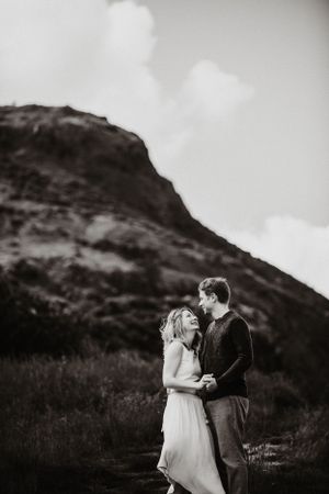 A couple shares a romantic moment against a dramatic mountain landscape in black and white photography.