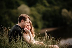 A romantic couple sits together in tall grass at sunset with soft natural lighting creating an intimate mood.
