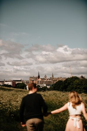 Two people stand on a hilltop overlooking Edinburgh's historic skyline with dramatic clouds in the distance.