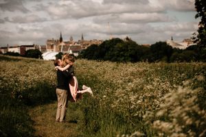 A couple embraces in a romantic moment amidst a wildflower meadow with a cityscape backdrop under moody clouds.