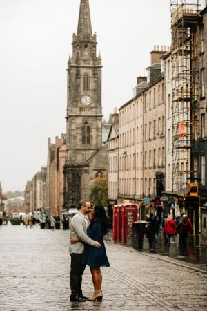 A couple embraces on the historic Royal Mile in Edinburgh with the Tron Kirk church spire and classic architecture in background.