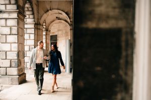 A couple walks together through a stone archway corridor while holding hands, dressed casually in daytime.