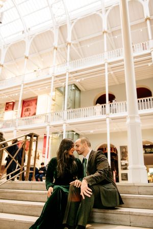 Elegant couple shares a romantic moment sitting on grand white marble steps inside a historic Victorian building.