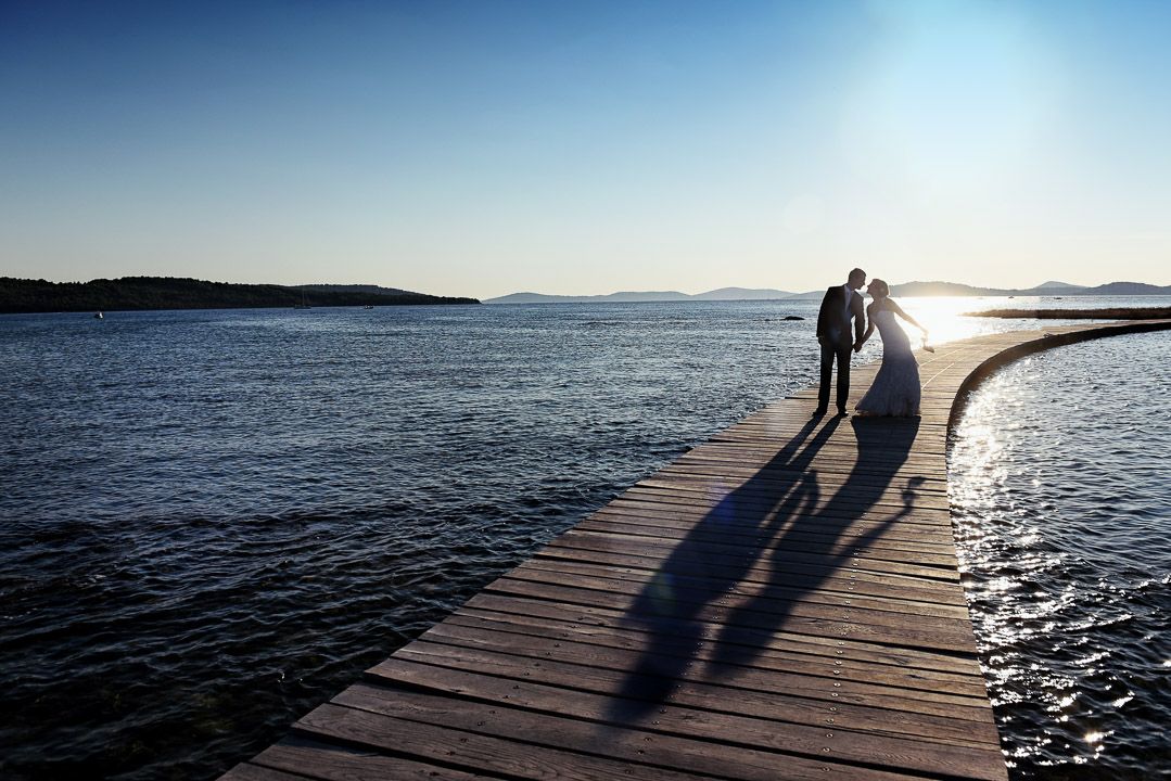 Two silhouettes cast long shadows on a wooden pier extending into calm waters at sunset with islands in the distance.