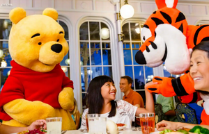 Costumed Disney characters including Winnie the Pooh and Tigger seated around a dining table at a restaurant event.