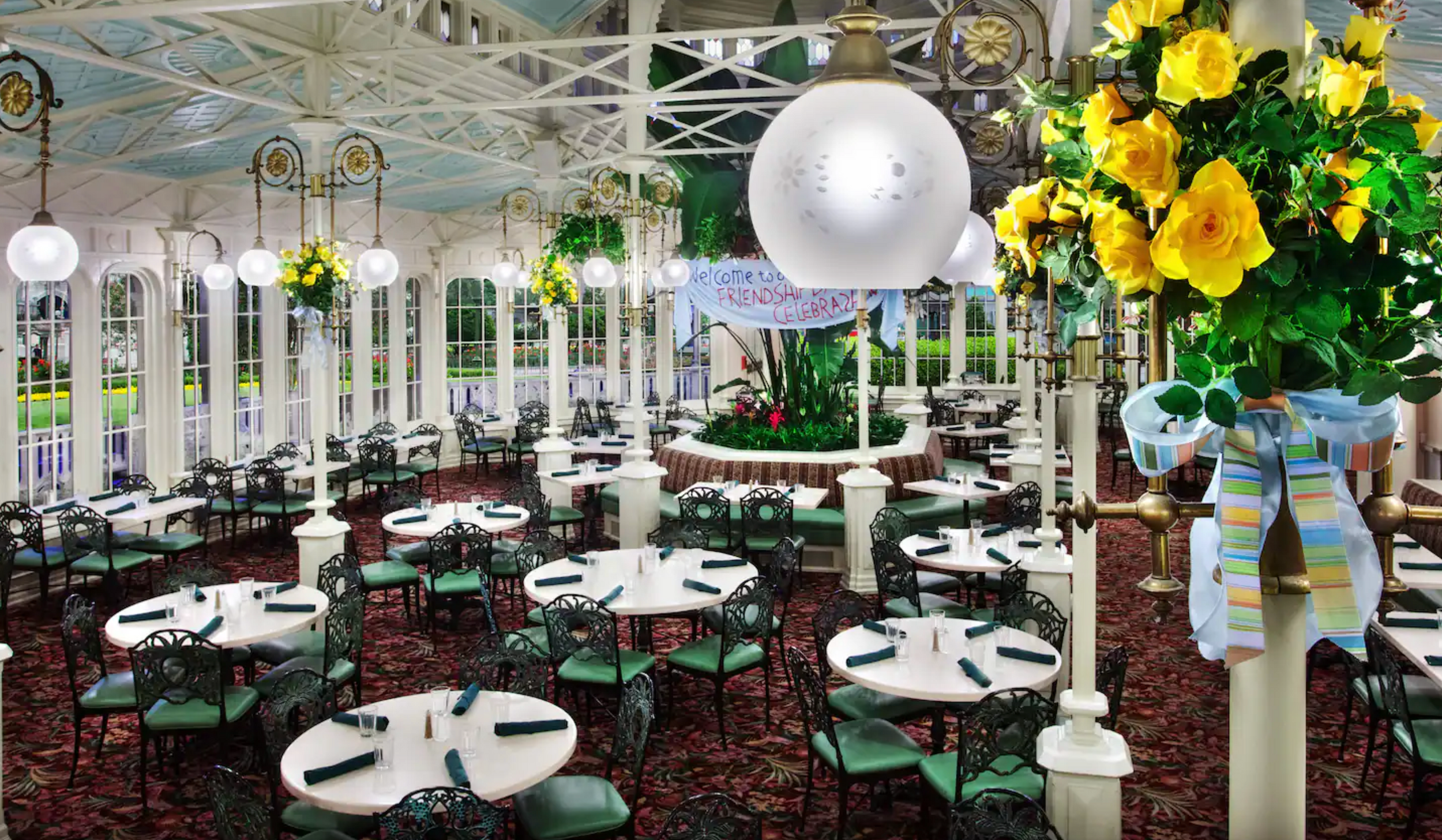 Elegant conservatory dining room with white round tables, green chairs, and yellow floral centerpieces under glass dome ceiling.
