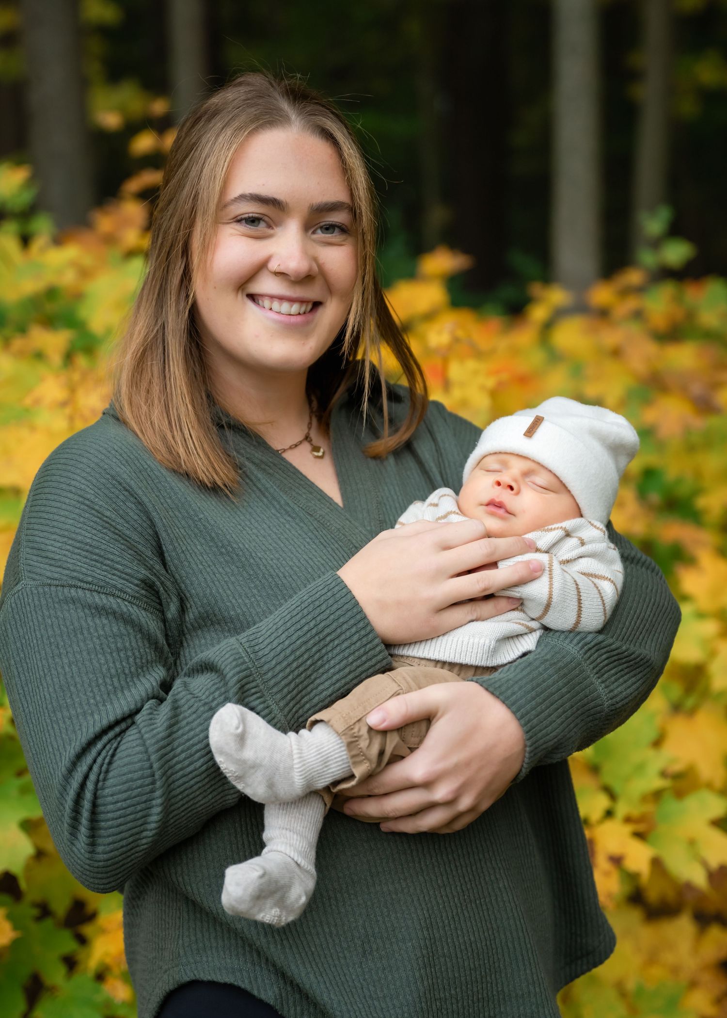 Smiling young woman in green sweater holds sleeping newborn baby outdoors with colorful autumn foliage background.