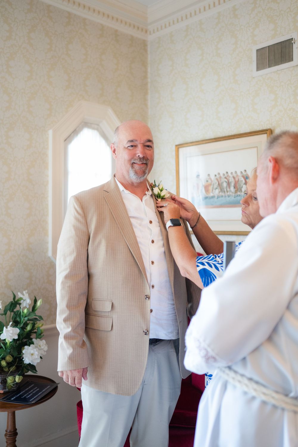 Groom in beige suit getting boutonniere pinned by another man in elegant wallpapered room before wedding ceremony.