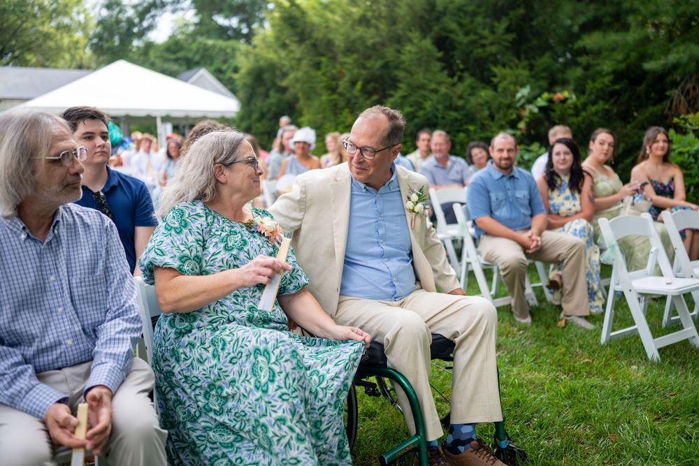 Outdoor wedding guests seated on white chairs, including a woman in a wheelchair, enjoying a garden ceremony.