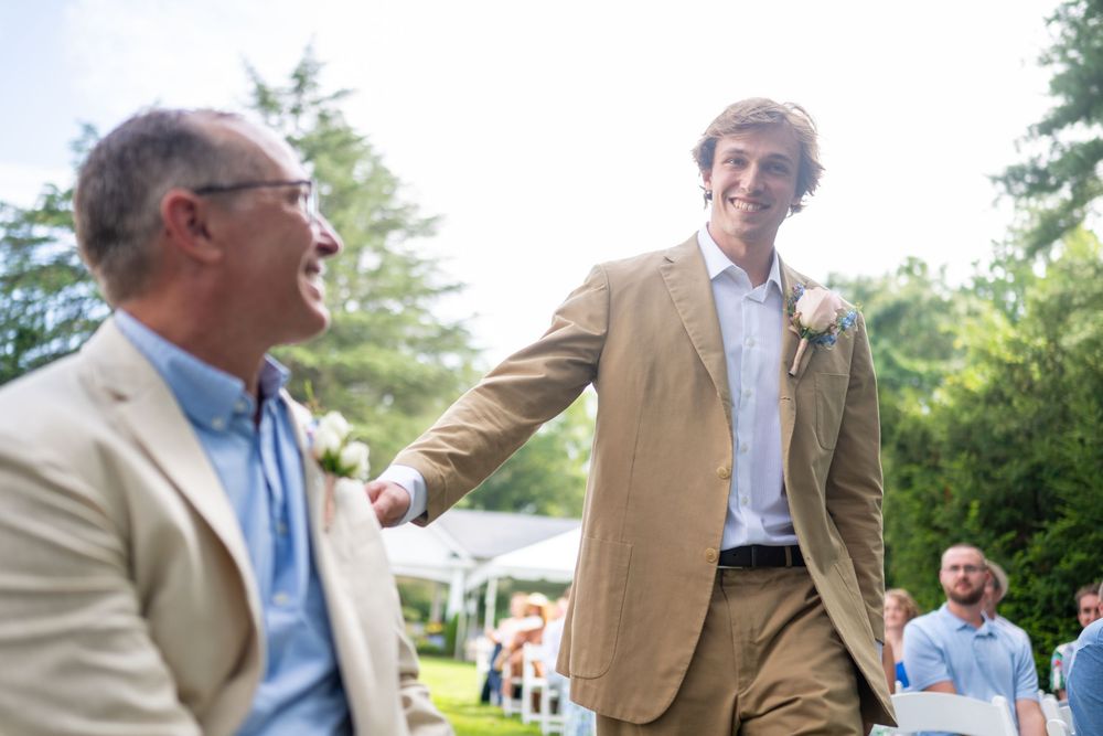 Two men in blazers laughing and shaking hands at an outdoor wedding celebration on a sunny day.