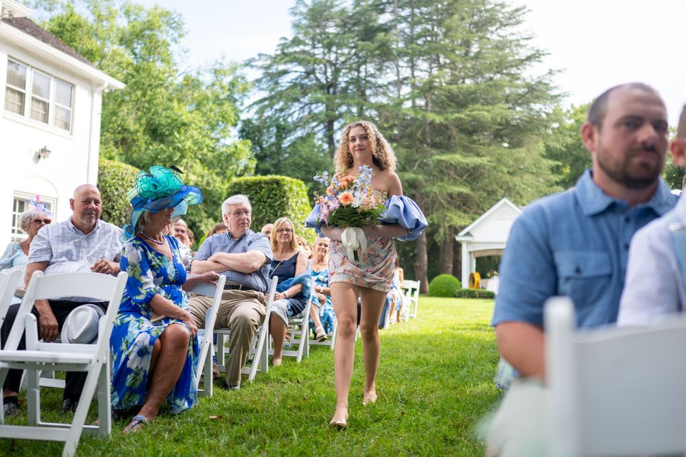 Barefoot bridesmaid walks down outdoor wedding aisle carrying flowers while guests seated in white chairs look on.
