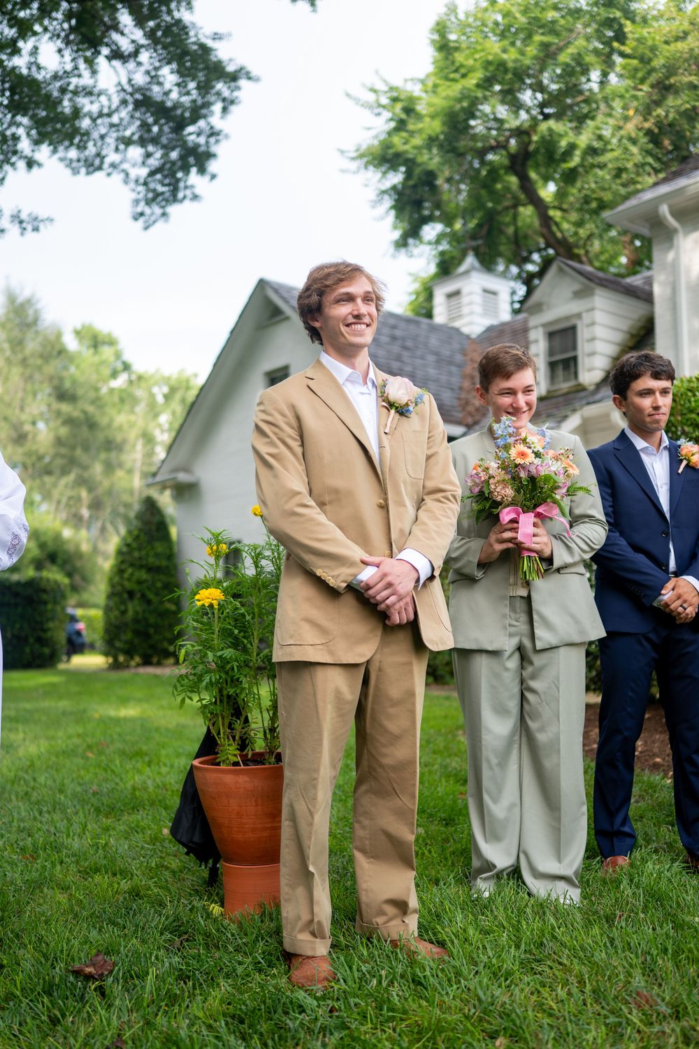 Smiling groom in tan suit stands at outdoor wedding ceremony on green lawn with wedding party behind him.