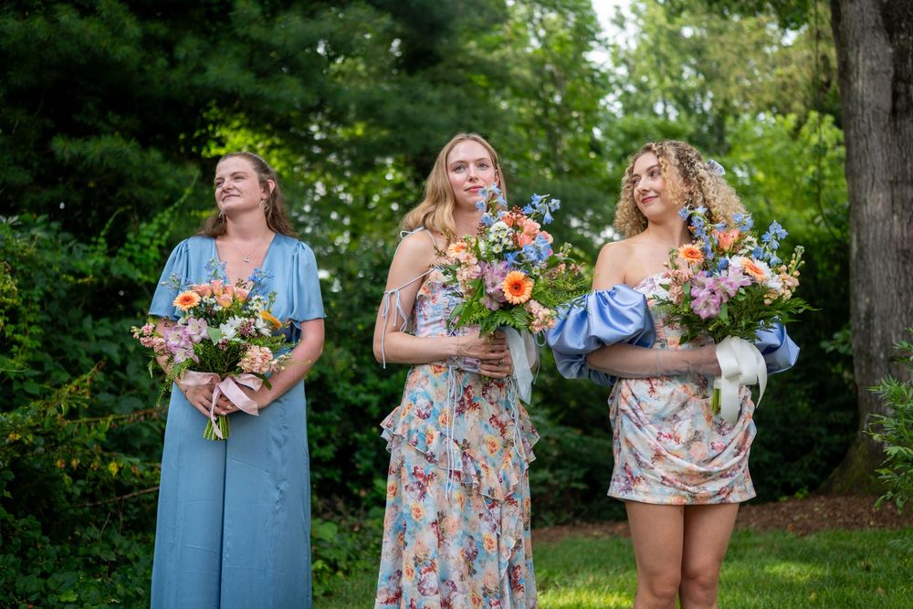Three bridesmaids in blue and floral dresses holding colorful bouquets of flowers outdoors at a wedding ceremony.