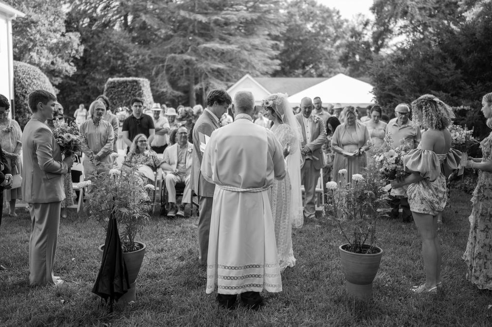 Black and white outdoor wedding ceremony with guests gathered on lawn, officiant in robes, bride in white dress, potted plants nearby.