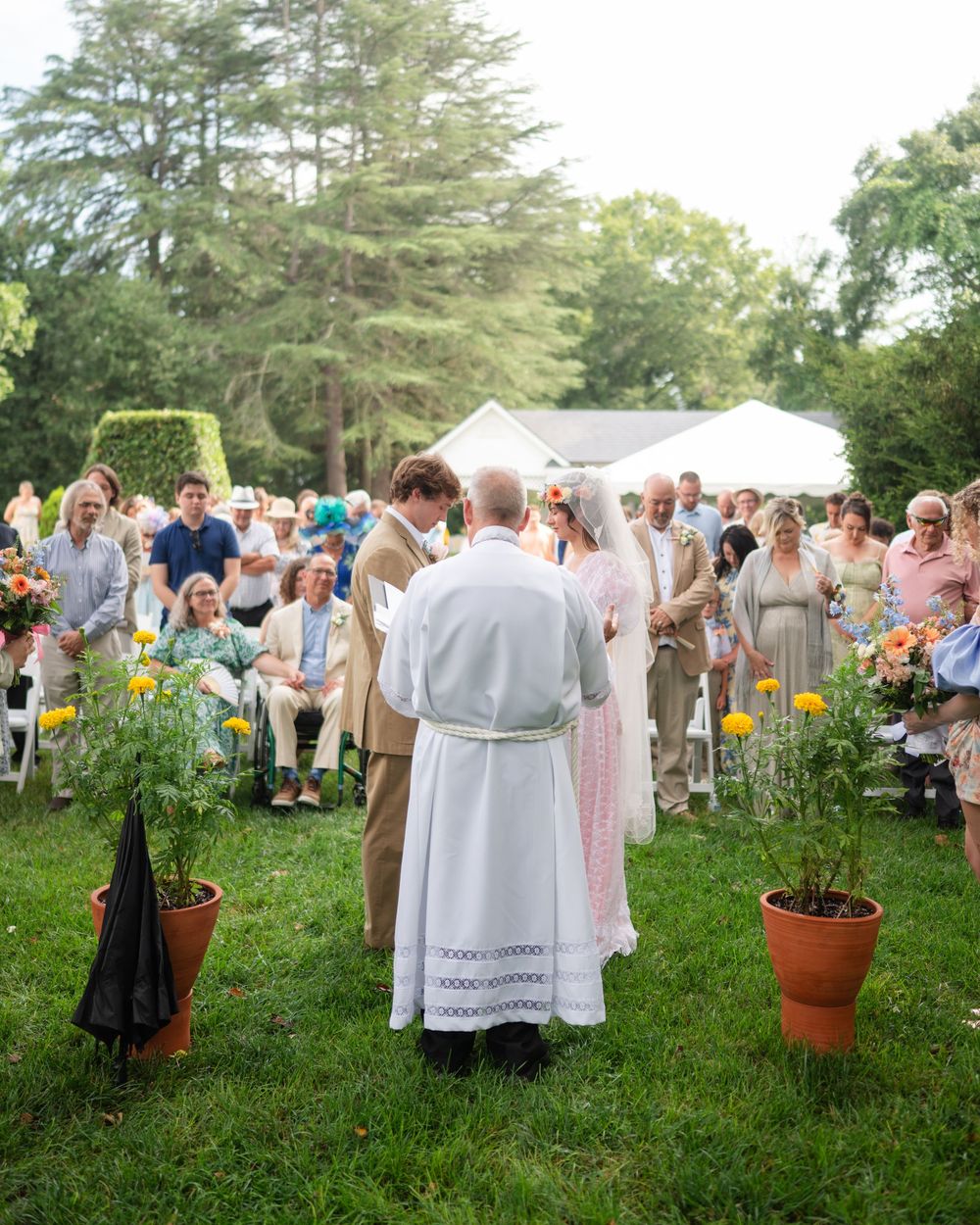 Outdoor wedding ceremony on green lawn with officiant, couple, and seated guests surrounded by trees and potted flowers.