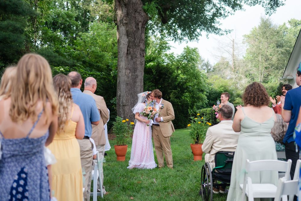 Bride in pink dress walks down grassy aisle toward groom at intimate outdoor backyard wedding ceremony.