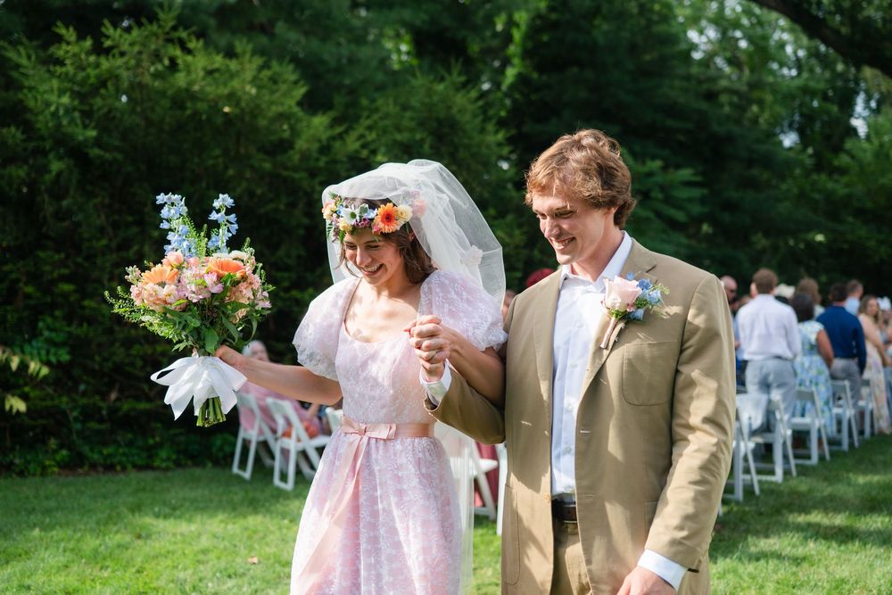 Bride in pink dress and floral veil walks with groom in tan suit at outdoor wedding ceremony.