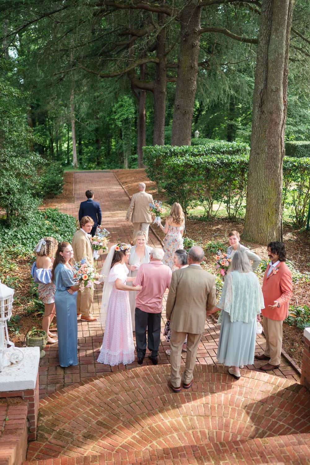 Aerial view of wedding guests gathered on brick patio surrounded by lush green trees during outdoor ceremony.