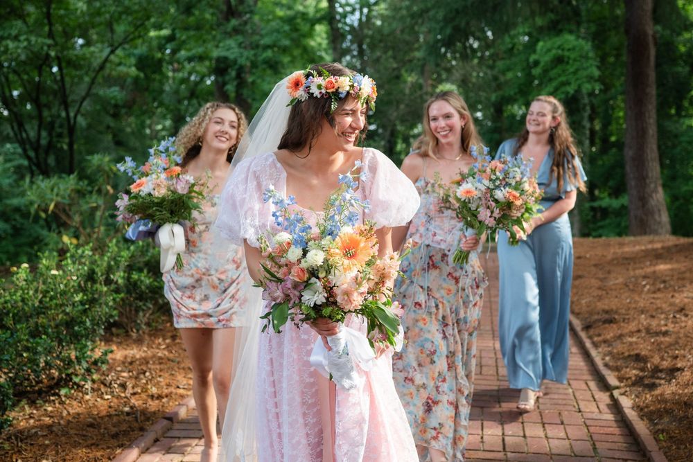 Bride in floral gown with flower crown walks with bridesmaids in blue dresses holding colorful bouquets outdoors.