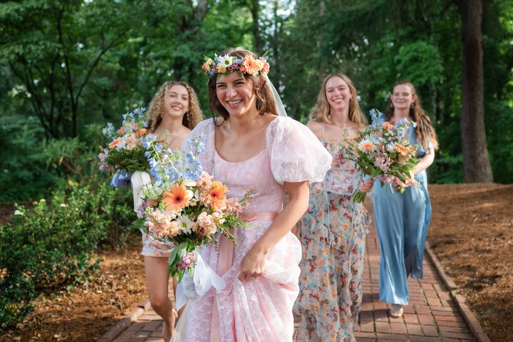 Bride in pink dress with floral crown leads bridesmaids holding colorful bouquets along a brick path in a lush garden.