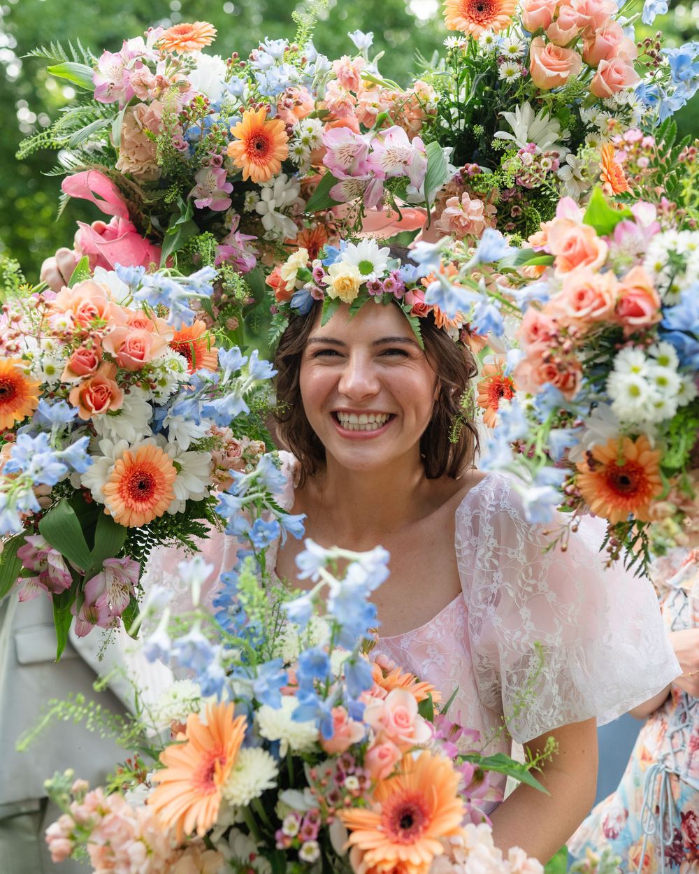Smiling bride surrounded by colorful floral arrangement with flower crown, featuring peach gerberas and blue blooms.