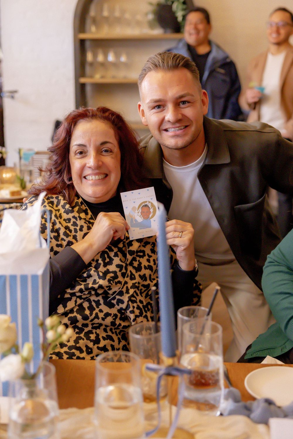Smiling woman in leopard print and man in leather jacket hold a small photo card at a festive table setting.