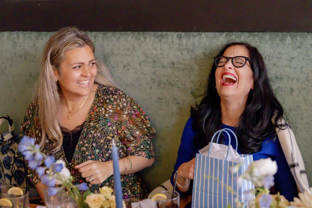 Two women laughing joyfully at a decorated table with flowers and gift bags at a social gathering.