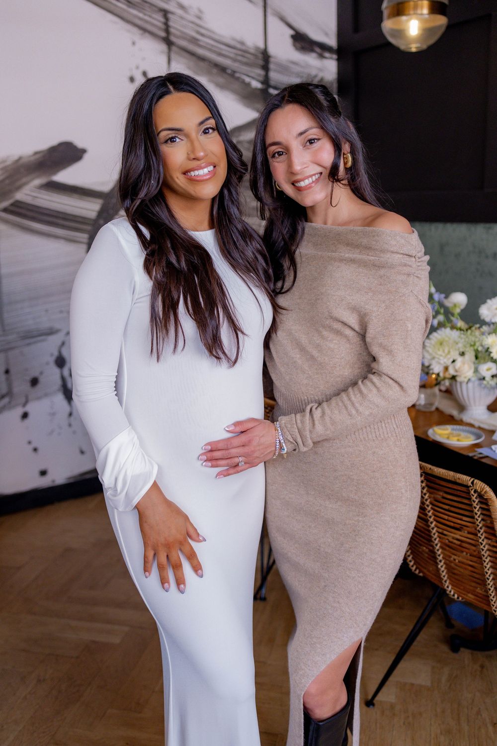 Two smiling women posing together at baby shower, pregnant woman in white dress with friend in beige.