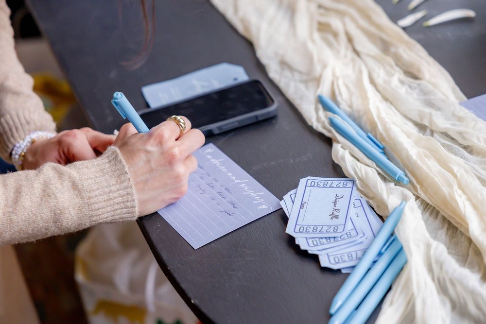 Woman writing on a notecard with a blue pen, surrounded by cards, pens, a phone, and white yarn on a dark table.