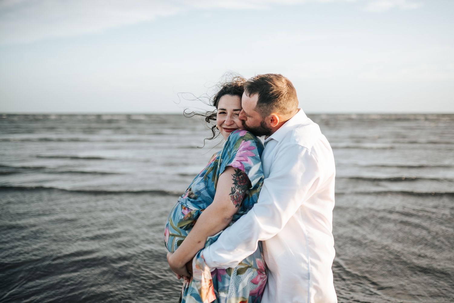Pregnant couple embracing and kissing on a beach with ocean waves, woman in floral dress laughing joyfully taken by Melbourne photographer.