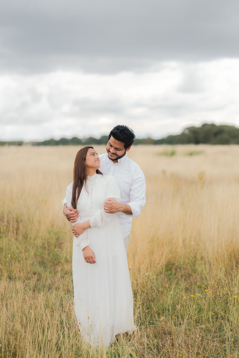 Couple in white outfits embracing in a golden field under cloudy skies.